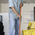 A janitor using a mop and bucket to wash the hallway floor of an office corridor in a Manhattan building.