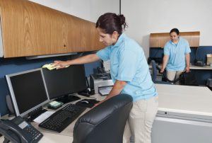 Two maids cleaning an office, one is vacuuming and the other is cleaning a computer monitor
