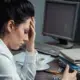 A stressed office worker holds her head in one hand and an empty stapler in the other, sitting at a messy and visibly dusty desk. Showcasing low productivity due to poor cleaning services.