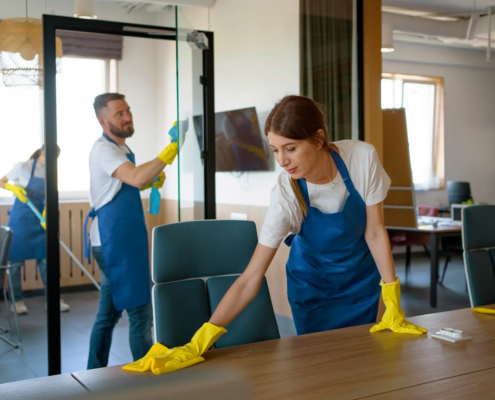 Commercial part-time janitorial services team in blue aprons cleaning modern office workspaces, wiping a desk and glass window with spray bottle.