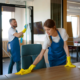 Commercial part-time janitorial services team in blue aprons cleaning modern office workspaces, wiping a desk and glass window with spray bottle.