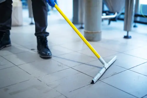 Close-up of a worker pushing dirty water with a floor squeegee on grey tiles, showcasing our thorough office floor deep cleaning service.