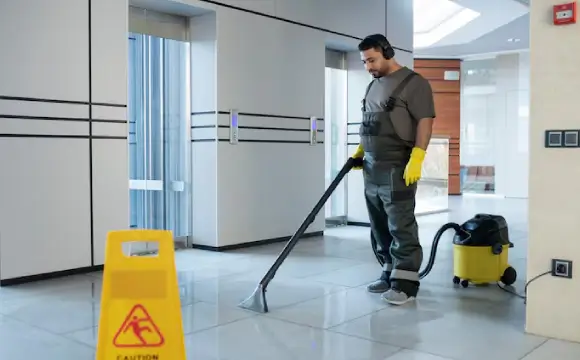 workspace-cleaning A commercial cleaner in grey overalls vacuums tiled floors near office elevators, representing flexible janitorial services for businesses in NYC.