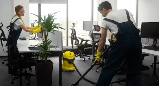 Two trained pro cleaners in navy overalls and yellow gloves dust workstations and vacuum around desk chairs in a carpeted office suite.