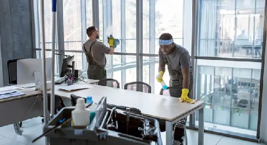 office-cleaning-contract Two professional cleaners for NYC businesses in gray coveralls and yellow gloves wipe a conference table and floor-to-ceiling windows in a New York City office.