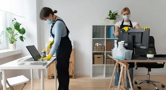 Two masked janitors in overalls and yellow gloves wipe down a laptop desk and a computer monitor in a small, well-organized workspace.