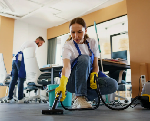 Two-person crew in blue aprons cleans floors and desks in an open office, showing what to expect from a commercial cleaning service provider in NYC.