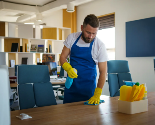 Cleaner in a blue apron and yellow gloves sprays and wipes a wooden conference table, part of routine work behind NYC office cleaning prices.