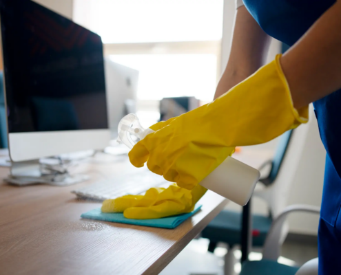 Cleaner in yellow rubber gloves wipes down a wooden desk near a keyboard and monitor, a simple way to improve office cleaning for NYC businesses.